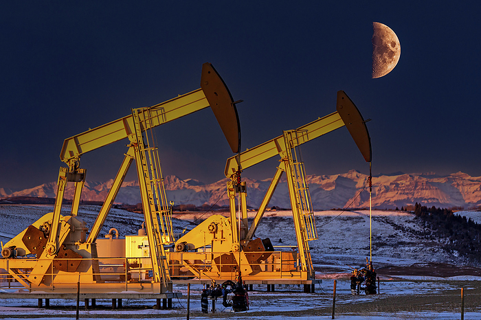 Canada Colourfully warm lit pumpjacks at sunrise in a snow covered field with glowing snow covered mountain range and half moon in a deep blue sky, West of Airdrie  Alberta, Canada, by Michael Interisano   Design Pics