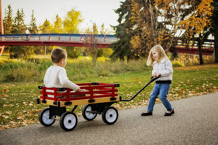 Young girl pulling her brother in a wagon at a city park along a river during the fall season; St. Albert, Alberta, Canada, by LJM Photo / Design Pics