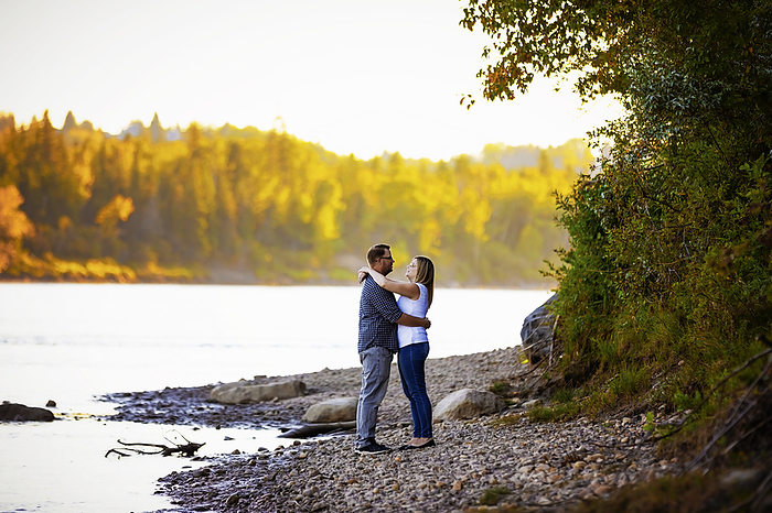 Married couple stand in an embrace beside a lake with autumn colours in the forest in the distance; Edmonton, Alberta, Canada, by LJM Photo / Design Pics