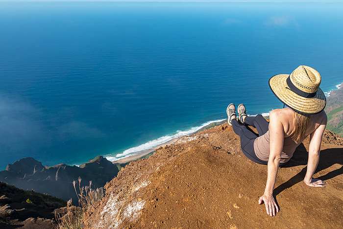 View taken from behind of a woman in a straw hat sitting on the top of a mountain cliff looking out onto the brilliant, blue water of the Pacific Ocean on the Kalalau Trail along the Napali Coast; Kauai, Hawaii, United States of America, by Living Moments Media / Design Pics