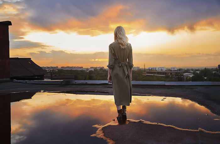 Blond young woman standing by puddle on rooftop under cloudy sky
