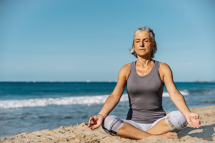 Mature woman sitting with legs crossed and meditating at beach
