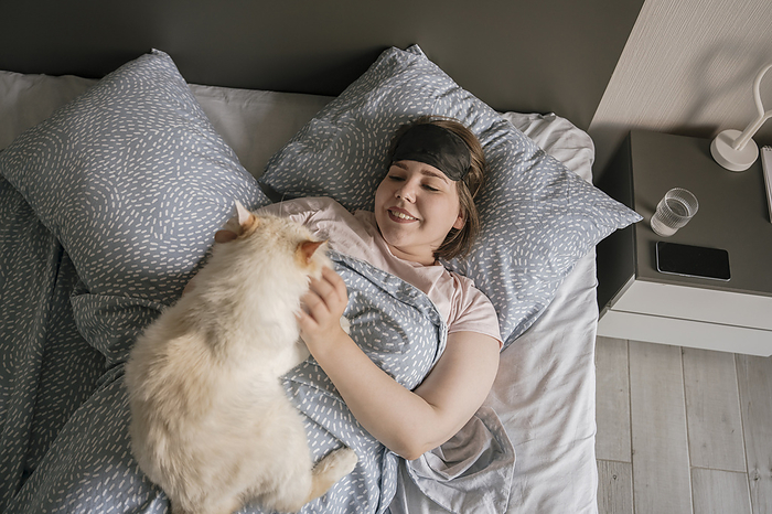 Smiling woman wearing eye mask stroking cat on bed