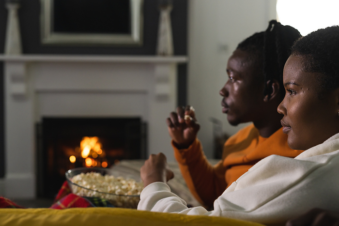 African american couple relaxing at home watching tv and eating popcorn. Relaxation, relationship, togetherness, communication, entertainment, winter, warmth and domestic life, unaltered.