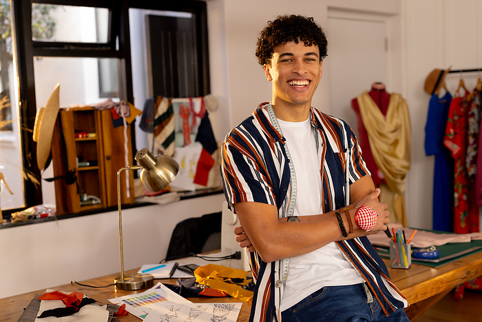 Focused biracial male fashion designer sitting on desk in studio and laughing. Fashion, design, creativity, clothing and small business, unaltered.