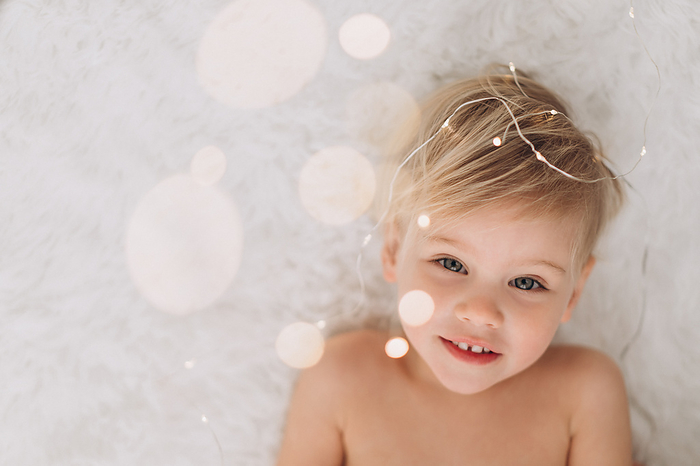 Naked child 2 years old lies and smiles at the lights from the garland, by Cavan Images / Liza Zavialova