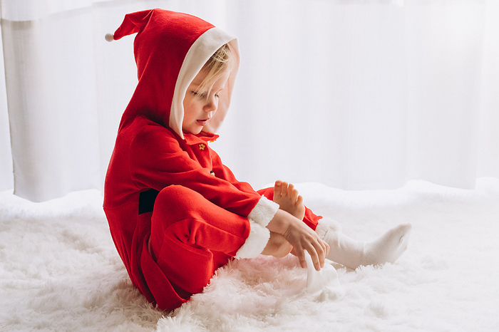 Child dressed as Santa sits and puts on socks, by Cavan Images / Liza Zavialova
