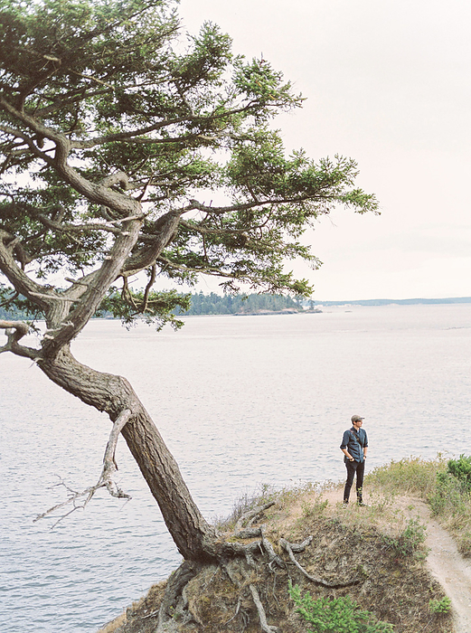 Man standing on overlook on the Pacific Northwest next to tree., by Cavan Images / Feather & Twine