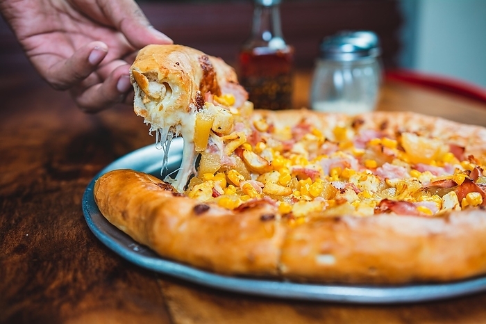 Hand taking a slice of ham and sweetcorn pizza. Homemade ham pizza with cheese and sweet corn served on wooden table, by Isai Hernandez
