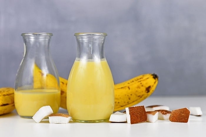 Two jars with yellow banana and coconut smoothie drink in jar next to ingredients, by Firn