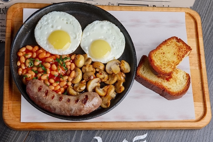 Cast iron skillet with fried eggs, sausage, mushroom and kidney beans with toasted bred on wooden board