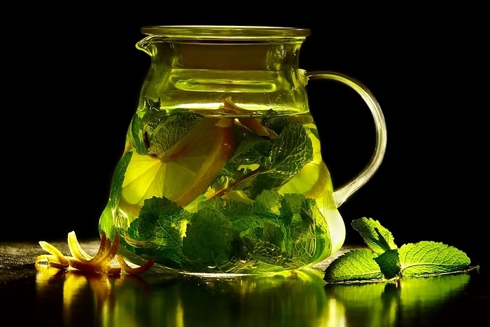 Transparent teapot of green tea with lemon and mint with ingredients. Jar of green tea with lime, lemon, mint on black slate stone board over black textured background