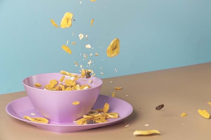 Healthy breakfast in pink bowl, banana chips, cornflakes, muesli flying, blue background, copy room