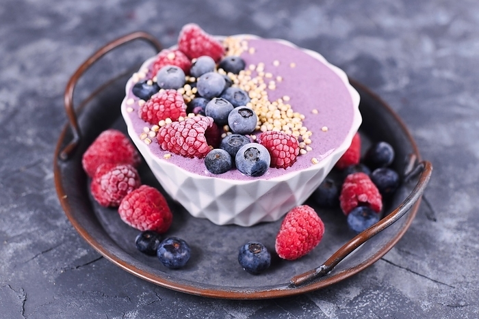 Healthy pink yogurt and fruit smoothie bowl decorated with raspberry, blueberry and puffed quinoa grain in white bowl on metal tray