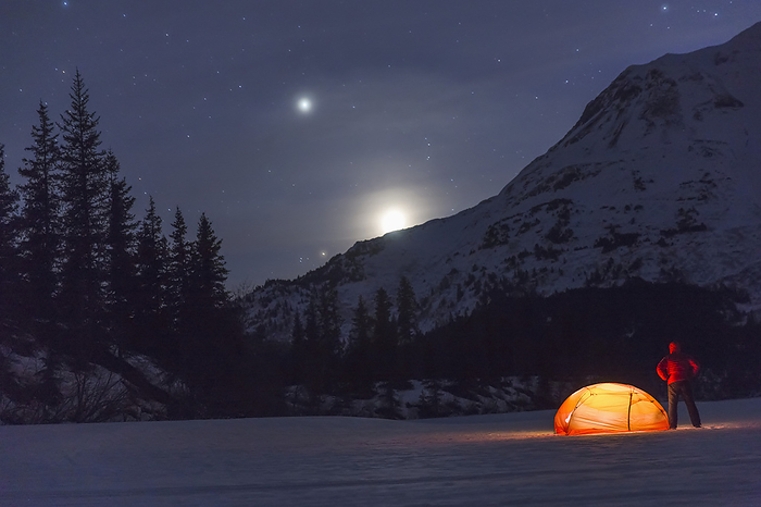 Nighttime View Of A Man Standing Next To A Lit Tent On A Snow Covered Trail Lake With The Moon, Stars, And Planets Overhead, Moose Pass, Kenai Peninsula, Southcentral Alaska, by Kevin G. Smith / Design Pics