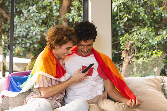 Happy diverse gay male couple covered with lgbt flag embracing and using smartphone at home. Domestic life, pride, technology and lifestyle, unaltered.