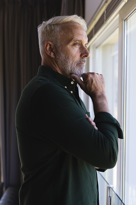 Thoughtful senior caucasian man looking through window at home. Senior lifestyle and domestic life, unaltered.