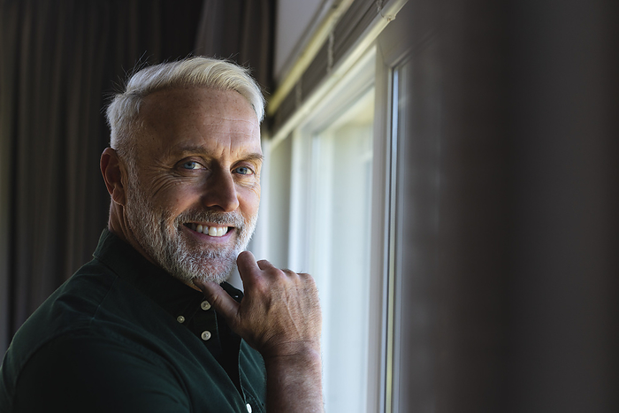 Portrait of happy senior caucasian man standing next to window at home, copy space. Senior lifestyle and domestic life, unaltered.