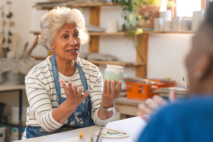 Happy biracial senior female potter with gray hair, discussing with others in pottery studio. Pottery, ceramics, handmade, local business, hobbies and craft, unaltered.