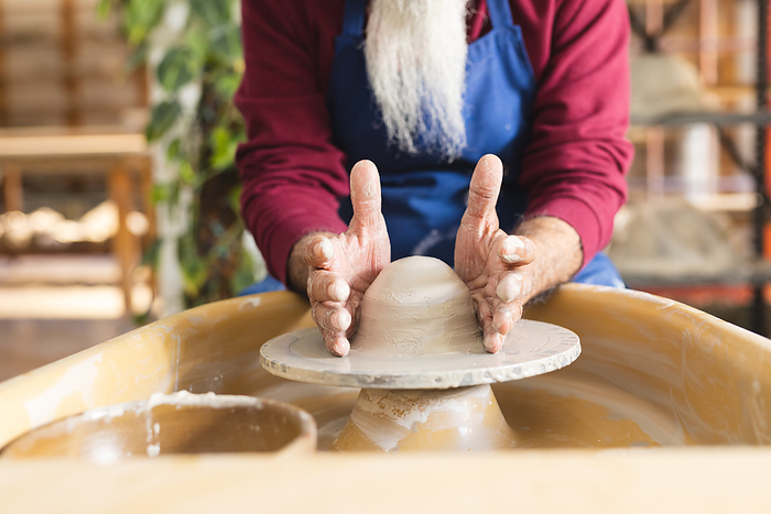 Focused biracial senior potter with long beard using potter's wheel in pottery studio. Pottery, ceramics, handmade, local business, hobbies and craft, unaltered.