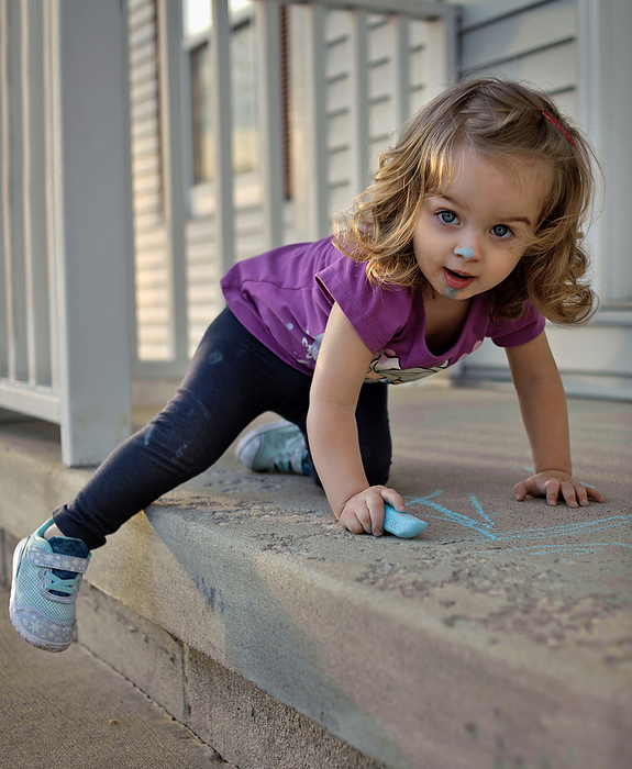 Little girl with blonde curls drawing with sidewalk chalk in summer, by Cavan Images / Joy Faith
