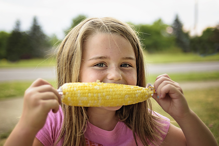Happy young girl eating yellow corn on the cob in summer, by Cavan Images / Joy Faith