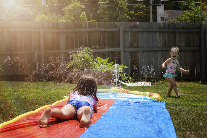 Sisters sliding down slip n  slide in summer, by Cavan Images / Joy Faith
