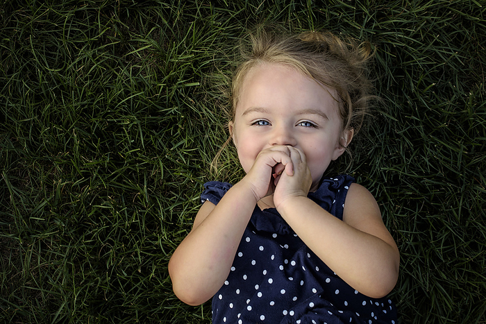 Beautiful little girl smiling while laying in the grass in summer, by Cavan Images / Joy Faith