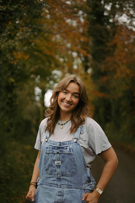 Young woman smiles in denim overall dungarees amidst fall forest, by Cavan Images / Sarah McKernan