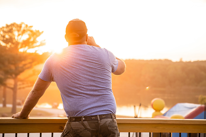 Man watching the sunset over lake while having a drink, by Cavan Images / Krista Taylor