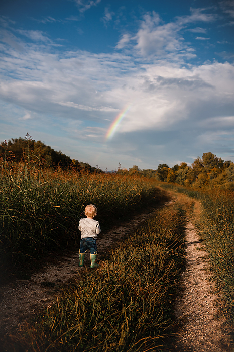 Young child on country road looking at rainbow on Autumn day, by Cavan Images / Krista Taylor