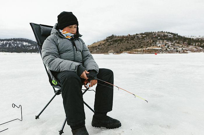 Teenage boy using fishing pole on frozen lake in winter, by Cavan Images / Anna Rasmussen Photographs