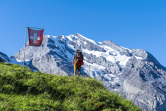 Rear view of a man with backpack hiking the Swiss Alps passing a Swiss flag flying, Oeschinensee, Kandersteg, Bern Canton, Switzerland, Europe Rear view of a man with backpack hiking the Swiss Alps passing a Swiss flag flying, Oeschinensee, Kandersteg, Bern Canton, Switzerland, Europe, by Paolo Graziosi