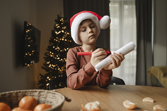 Boy wearing santa hat writing on paper at table in home