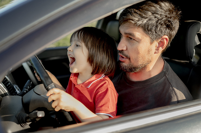 Cheerful boy holding steering wheel with father sitting in car