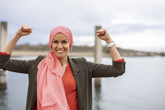 Smiling woman wearing headscarf flexing muscles by sea