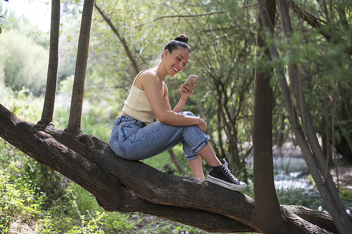 Happy woman using smart phone sitting on tree in forest