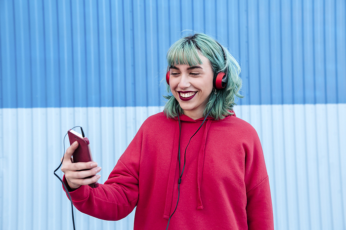 Portrait of laughing young woman with blue dyed hair with headphones taking selfie with mobile phone
