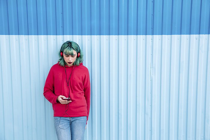 Young woman with blue dyed hair listening music with headphones while looking at smartphone