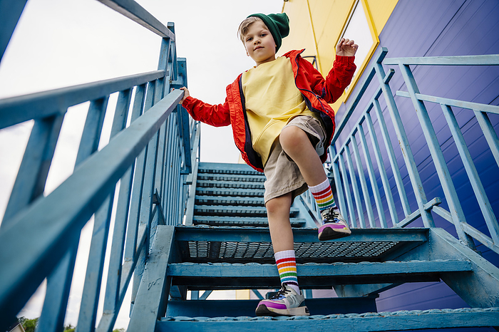 Boy in red jacket standing on one leg on metallic staircase by building