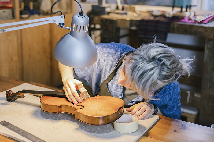 Young luthier working at her workshop Instrument maker arranging bridge part on violin at desk in workshop