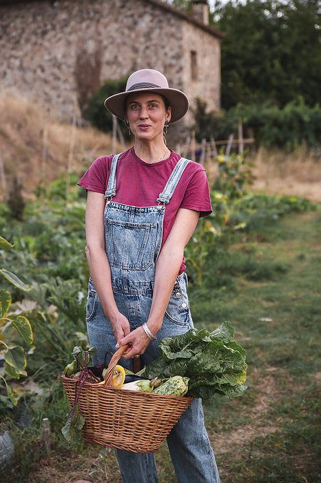 Smiling woman holding wicker basket with vegetables in orchard