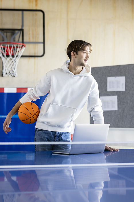 Young trainee standing with basketball and laptop in office