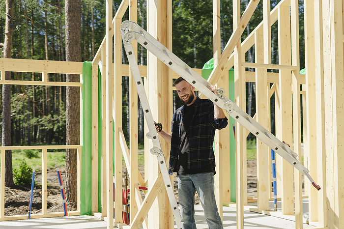 Construction team on a new site in the woods, working with tools, communicating, checking engineering plans Smiling engineer standing near wooden frame on sunny day