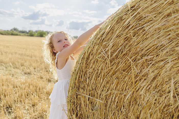 Blond girl touching bale of straw in field on sunny day