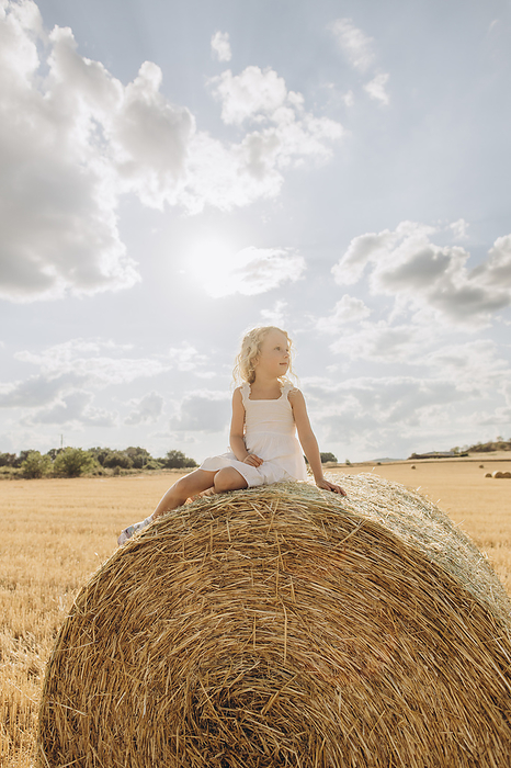 Contemplative blond girl sitting on bale of straw in field on sunny day