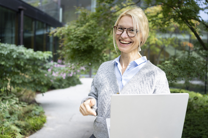 Smiling businesswoman wearing eyeglasses holding laptop at office park