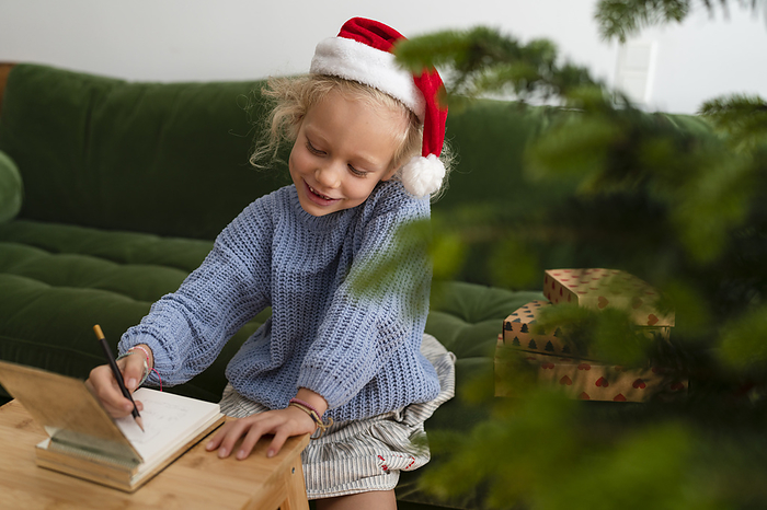 Cheerful girl writing letter to Santa and sitting on sofa at home