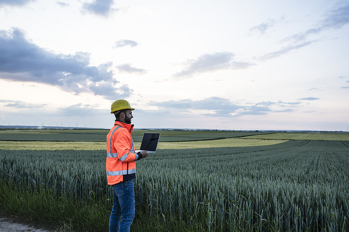 Engineer in reflective clothing holding laptop looking at crop in field