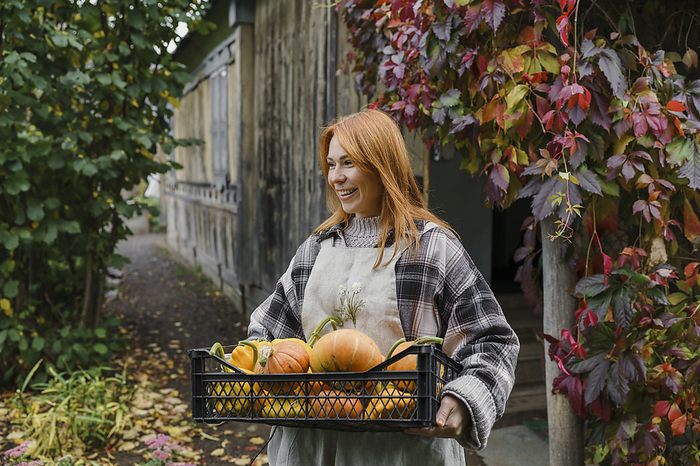 Happy redhead farmer carrying crate of pumpkins at farm